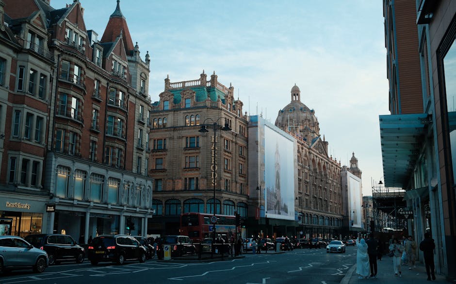A city street scene featuring historic and modern architecture with a large ornate building in the background topped with a decorative dome and turquoise accents. The street is lined with multi-story buildings constructed from red brick and stone, with numerous windows and intricate facades. Several parked cars, including a red double-decker bus, are visible along the curb. Pedestrians, some dressed in formal attire, are walking along the sidewalk, and a few are engaged in conversation. On the right side, a contemporary glass-fronted retail store with a blue awning is partially visible. The sky is clear with soft daylight illuminating the scene, emphasizing the architectural details. The overall setting appears to be in a busy urban district, typical of a central commercial or shopping area, consistent with independent or private waste handling practices often seen in city centres.