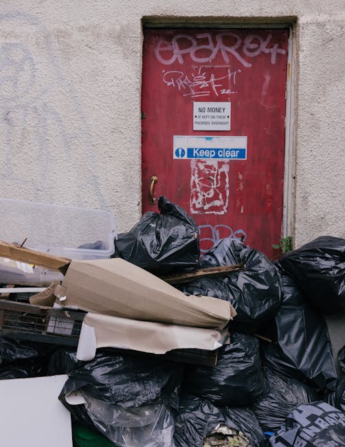 A collection of black bin bags, cardboard boxes, and discarded packaging piled up in front of a weathered red metal door on an exterior wall, with graffiti and signs including a 'Keep Clear' notice. The bags appear to be filled with household waste, with some loose items like cardboard and foam packaging resting atop or leaning against the pile. The wall around the door has a rough, light-colored textured surface, and there is minimal surrounding environment visible, indicating an urban setting. The scene reflects an instance of waste accumulation awaiting collection, suggestive of independent rubbish removal or private disposal, aligning with services offered by Rubbish Removal Knightsbridge for non-standard waste clearance. The lighting is natural, with soft shadows, emphasizing the practical nature of the scene. This detailed description ensures clear understanding of the visible objects, materials, and environmental context, supporting accessibility and relevance to rubbish removal service content.