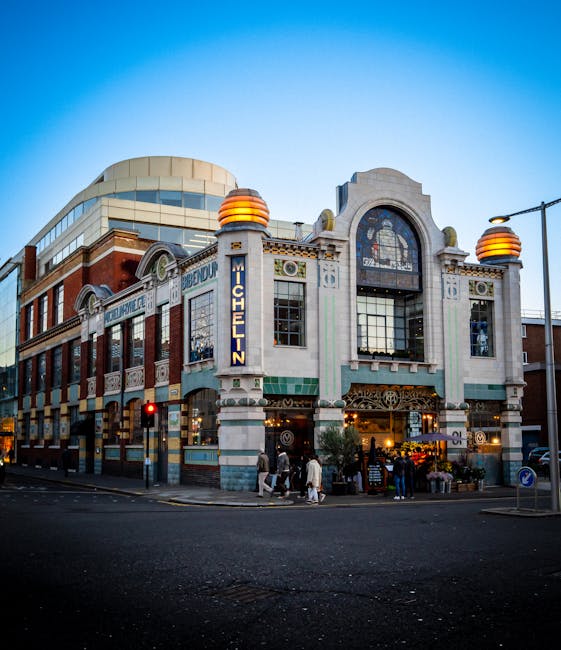 A multi-story, historic-style building with intricate architectural details, including arched windows and decorative stonework, situated at a street corner during early evening with a clear blue sky. The facade features a prominent vertical sign displaying the name 'Michelin' in blue illuminated letters. The ground level has large glass doors and windows, with some greenery and outdoor plants visible near the entrance. Surrounding the building are modern adjacent structures, streetlights, a traffic light showing red, and pedestrians crossing the road. The scene captures urban activity, with people walking along the pavement and city-life atmosphere. The lighting suggests a transition from daylight to dusk, highlighting the building’s distinctive features and the street’s vibrancy, typical of a busy commercial area in Knightsbridge. This setting subtly aligns with themes of urban development, commercial heritage, and independent city services, such as private waste collection and rubbish removal, though no waste-related objects are visible in the image.
