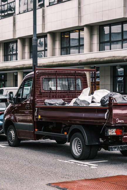 A small, dark red flatbed truck parked on a city street in front of a modern office building with large glass windows and white concrete exterior. The truck's cargo bed is loaded with various types of rubbish, including large black and white bags, and other miscellaneous waste materials. The truck features a metal mesh safety screen behind the cab and has a single rear axle with small wheels. The scene is illuminated by natural daylight, and the surrounding environment includes parked vehicles and a partially visible pedestrian crossing marking on the road, indicating an urban setting suitable for private waste removal services such as those provided by Rubbish Removal Knightsbridge, assisting in alternative rubbish collection solutions.