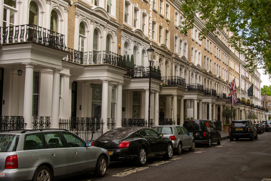 The image depicts a row of elegant, multi-storey white terraced buildings with classical architectural features, including decorative columns, ornate black wrought iron balconies, and large sash windows. The buildings are situated along a quiet urban street with parked cars lining the curb, including a silver station wagon, a black luxury vehicle, and a grey compact car. The street incorporates a narrow sidewalk with a black iron fence separating the pavement from the building entrances. On the right side of the image, Union Jack and other flags hang from flagpoles attached to the buildings, and a traditional black lamppost stands among lush green trees with dense foliage, partially obscuring the upper floors. The scene is evenly lit with natural daylight, suggesting a clear day with some cloud cover, and the overall atmosphere conveys a well-maintained residential area. This setting is typical of London’s upscale neighborhoods, where private property and alternative waste management options may be used for rubbish collection, exemplified by the neat arrangement of vehicles and the maintained façade of the buildings, consistent with professional rubbish removal services such as those offered by Rubbish Removal Knightsbridge.