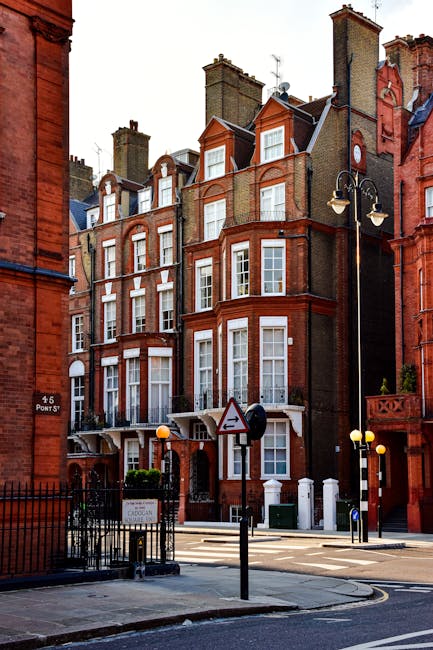 The image depicts a row of elegant, multi-storey white terraced buildings with classical architectural features, including decorative columns, ornate black wrought iron balconies, and large sash windows. The buildings are situated along a quiet urban street with parked cars lining the curb, including a silver station wagon, a black luxury vehicle, and a grey compact car. The street incorporates a narrow sidewalk with a black iron fence separating the pavement from the building entrances. On the right side of the image, Union Jack and other flags hang from flagpoles attached to the buildings, and a traditional black lamppost stands among lush green trees with dense foliage, partially obscuring the upper floors. The scene is evenly lit with natural daylight, suggesting a clear day with some cloud cover, and the overall atmosphere conveys a well-maintained residential area. This setting is typical of London’s upscale neighborhoods, where private property and alternative waste management options may be used for rubbish collection, exemplified by the neat arrangement of vehicles and the maintained façade of the buildings, consistent with professional rubbish removal services such as those offered by Rubbish Removal Knightsbridge.