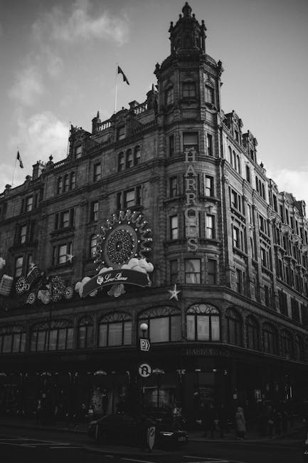 A black-and-white image of a historic multi-storey building situated at a busy street corner. The building features ornate architectural details, including decorative cornices, columns, and window surrounds typical of early 20th-century design. The facade is made of stone or brick with a textured appearance, and several windows are visible on each level, some with arched tops. At the center of the building, a vertical sign spells out 'HARRODS' in illuminated letters. On the ground floor, there are commercial storefronts with large display windows, and above these, a decorative sign indicates a pâtisserie or restaurant, with a small globe and star ornamentation. Flags are mounted on the upper corners of the building, fluttering in the breeze. The street below is bustling with pedestrians and vehicles, and there is a parking sign visible near the curb. The overall scene reflects an iconic, urban setting associated with private retail spaces and independent collection of waste, typical in a city environment where rubbish removal services such as those offered by Rubbish Removal Knightsbridge are important for maintaining cleanliness around prominent commercial landmarks.
