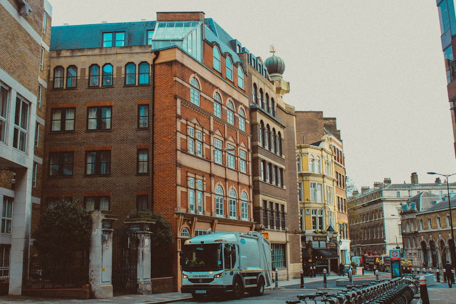 A street scene in an urban environment featuring a variety of multi-story buildings with architectural details including arched and rectangular windows, some with decorative trim, and a distinctive copper-domed tower visible on one building. The facades display a mix of brickwork and painted finishes in earthy and pastel tones. In the foreground, a light blue waste collection truck with a compact, modern design is parked along the kerb, positioned in front of a gated entrance with stone pillars and greenery surrounding it. The street surface appears to be asphalt, lined with black bollards and bicycles locked in racks on the right side. A few pedestrians and additional vehicles are visible further down the street, with overcast lighting casting a neutral tone over the scene, reflecting a typical city environment that might require private waste removal services. Rubbish Removal Knightsbridge's visual context is subtly represented by the presence of waste collection infrastructure, indicating on-site clearance or alternative waste handling activities typical in urban rubbish removal operations.