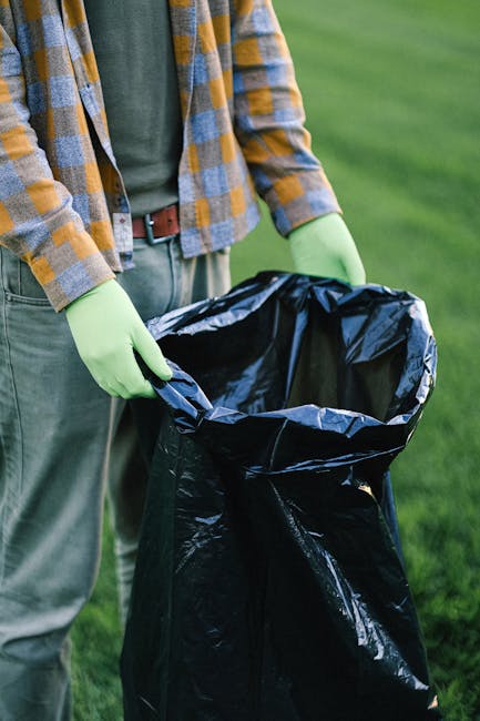 A person wearing a green long-sleeved shirt, a yellow and grey checkered jacket, and light grey trousers is standing outdoors on a grassy area. They are holding open a large black garbage bag lined with a shiny, reflective material. The person's hands are covered with light green disposable gloves, and they are preparing to dispose of waste into the bag. The background shows a well-maintained lawn, suggesting a clean and tidy environment. This scene depicts an individual involved in waste collection or rubbish removal, aligning with private or independent waste handling services. The image emphasizes proper waste disposal methods, with attention to detail in the protective gloves and the large capacity of the bag suitable for rubbish collection, possibly related to professional rubbish removal activities that avoid reliance on local authority collection, consistent with services offered by Rubbish Removal Knightsbridge.