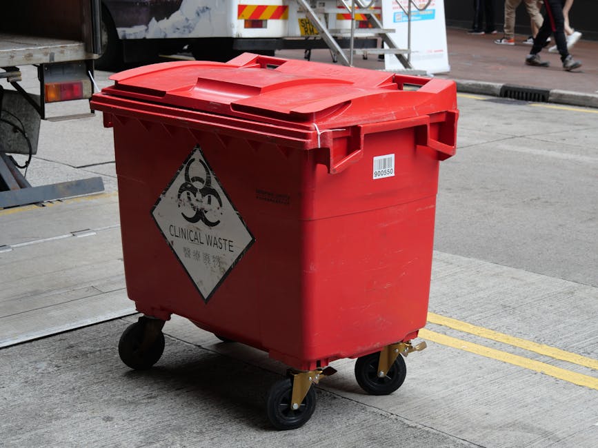 A red wheeled bin with a hinged lid, marked with a black and white biohazard symbol and the words 'Clinical Waste' in both English and Chinese, positioned on a concrete sidewalk. The bin appears to be made of durable plastic with a textured surface and is situated near the edge of the pavement, adjacent to a road with visible double yellow lines. In the background, partially visible, are various elements including a vehicle with a reflective white and yellow safety pattern, metal stairs leading up to a platform, and pedestrians walking on the sidewalk. The lighting suggests daytime with natural light illuminating the scene, and the setting indicates a designated area for waste disposal, aligning with private or specific waste removal services like those provided by Rubbish Removal Knightsbridge, specializing in rubbish collection and waste management outside standard local authority collections.
