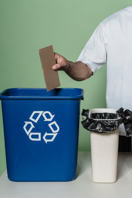 A person wearing a white shirt is holding a rectangular piece of cardboard above a blue recycling bin with a white recycling symbol on the front, positioned on a light-colored surface against a pale green background. To the right of the blue bin, there is a smaller white bin lined with a black plastic bag, which is partially filled with waste. Additional black plastic bags are visible next to it, resting on the surface. The scene depicts an organized approach to waste segregation, relevant to private rubbish collection and on-site waste disposal practices that Rubbish Removal Knightsbridge may facilitate, illustrating the separation of recyclable materials from general waste in a professional setting.