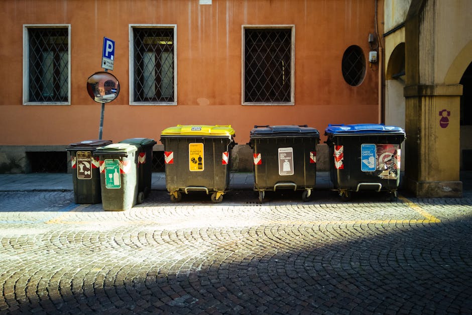 A red wheeled bin with a hinged lid, marked with a black and white biohazard symbol and the words 'Clinical Waste' in both English and Chinese, positioned on a concrete sidewalk. The bin appears to be made of durable plastic with a textured surface and is situated near the edge of the pavement, adjacent to a road with visible double yellow lines. In the background, partially visible, are various elements including a vehicle with a reflective white and yellow safety pattern, metal stairs leading up to a platform, and pedestrians walking on the sidewalk. The lighting suggests daytime with natural light illuminating the scene, and the setting indicates a designated area for waste disposal, aligning with private or specific waste removal services like those provided by Rubbish Removal Knightsbridge, specializing in rubbish collection and waste management outside standard local authority collections.
