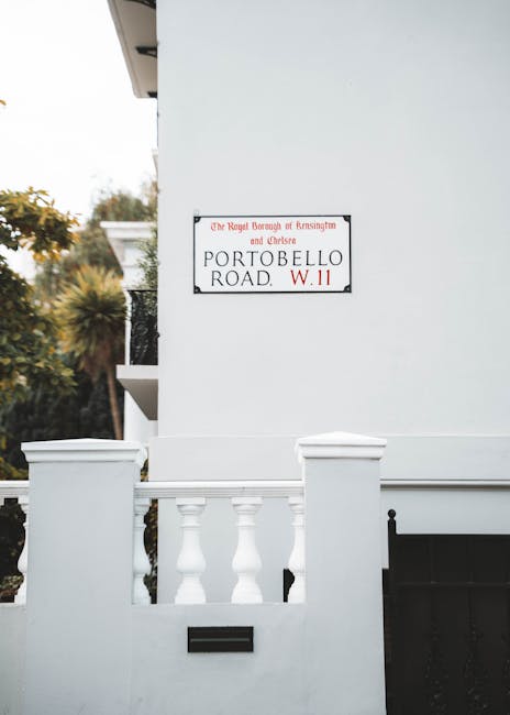 A white residential building with a small balcony enclosed by white balustrades is visible in the foreground, featuring a black mailbox inset into the wall below the balcony. Mounted on the building’s exterior wall is a rectangular street sign with a white background and red and black text, indicating the address as 'Portobello Road, W11,' and mentioning the Royal Borough of Kensington and Chelsea. In the background, there are blurred trees and foliage, suggesting a suburban or urban environment with greenery. The scene is lit with natural daylight, highlighting the clean, smooth finish of the white walls and the detailed architectural elements of the balustrade and sign. The overall setting appears tidy and well-maintained, and the presence of the sign indicates an official street designation, relevant to local waste disposal and independent collection services such as those offered by Rubbish Removal Knightsbridge, which specialize in alternative waste handling methods for properties in the area.