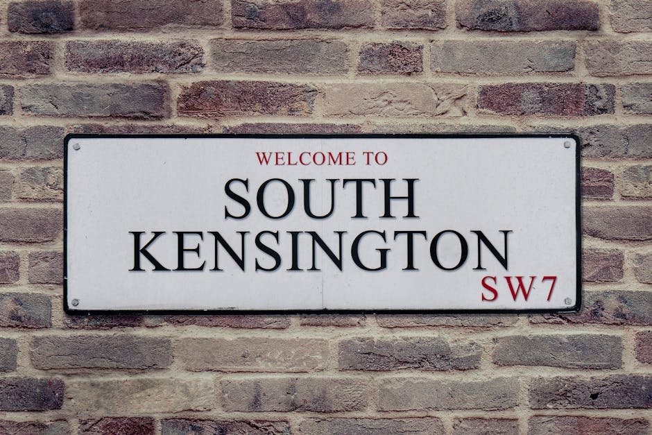 A rectangular white metal street sign mounted on a brick wall, displaying the words 'Welcome to South Kensington' in red and black capital letters, with the postcode 'SW7' also in red at the bottom right corner. The brick wall behind the sign consists of reddish-brown bricks with visible mortar joints, creating a textured background. The lighting is natural, illuminating the sign evenly and highlighting the contrast between the smooth surface of the sign and the rough texture of the brick wall. This image relates to the context of rubbish removal and property management in South Kensington, an area where private site clearance may be used to handle bulky waste following local council rules, reflecting the importance of proper disposal practices and independent collection services like those offered by Rubbish Removal Knightsbridge.