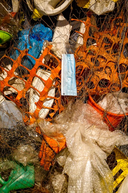 A close-up view of a tightly packed collection of mixed waste materials entangled in a fine netting, including orange plastic crates, crumpled plastic bags, and various discarded items. In the center, a light blue disposable face mask hangs amidst the clutter. Surrounding objects feature different textures such as smooth plastic, crinkled film, and rough surfaces, with colors predominantly in shades of orange, white, green, and yellow. The waste appears to be placed on a flat surface, possibly outdoors, with no clear environmental context visible. The scene emphasizes refuse that may be slated for disposal or collection by independent waste management services such as Rubbish Removal Knightsbridge, illustrating the diverse nature of refuse that requires specialist removal or disposal. The lighting is neutral, without strong shadows, and the overall composition highlights the tangled, disorganized state of the waste material, aligning with themes of private waste handling and alternative rubbish collection solutions.