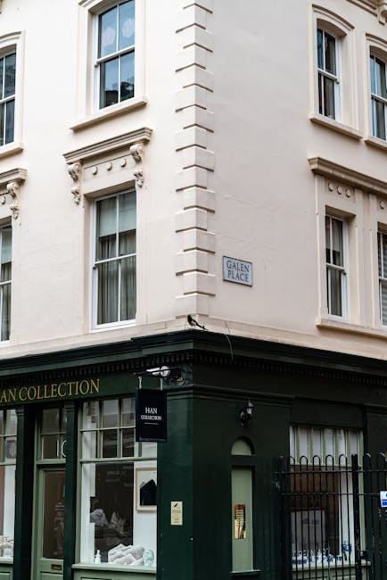 The image shows a corner of a historic, three-story building with a pale cream-colored façade and tall sash windows with white frames and decorative molding. The ground level features a dark green shopfront with large glass windows and a door, with a sign above reading 'HAN COLLECTION.' A small black square sign attached to the building displays the name 'HAN COLLECTION,' and a security camera is mounted above the storefront. A narrow street sign indicates the location as 'Galen Place,' positioned on the building’s corner. The environment appears urban, with the building situated on a quiet street. The overall scene is lit by natural daylight, with mild shadows indicating a partly cloudy sky. The style and setting suggest an area where independent retail or service businesses operate, often requiring occasional rubbish removal or private waste handling, which can be managed by services like Rubbish Removal Knightsbridge, supporting cleaner and more efficient waste disposal consistent with local regulatory standards.