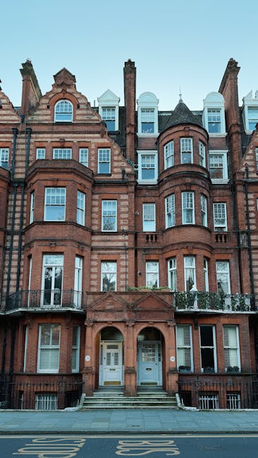The image depicts a multi-storey Victorian-style brick residential building with red-brown masonry, ornate architectural details, and numerous sash windows. The facade features distinct bay windows on each level, some with metal railings and flowerpots, and a central entrance with steps leading up to white double doors set beneath an arched brick surround. The building's roof includes chimney stacks, dormer windows, and decorative gables, with some sections finished in white or grey materials. The pavement in front shows a bus stop marking on the street, indicating an urban setting. The clear sky in the background suggests daytime, and the overall scene reflects a typical residential front, where private rubbish collection services, such as those offered by Rubbish Removal Knightsbridge, are often employed for on-site clearance or waste disposal without reliance on local authority collection. This detailed visual context supports the importance of professional rubbish removal for maintaining property tidiness and managing waste efficiently in residential areas.