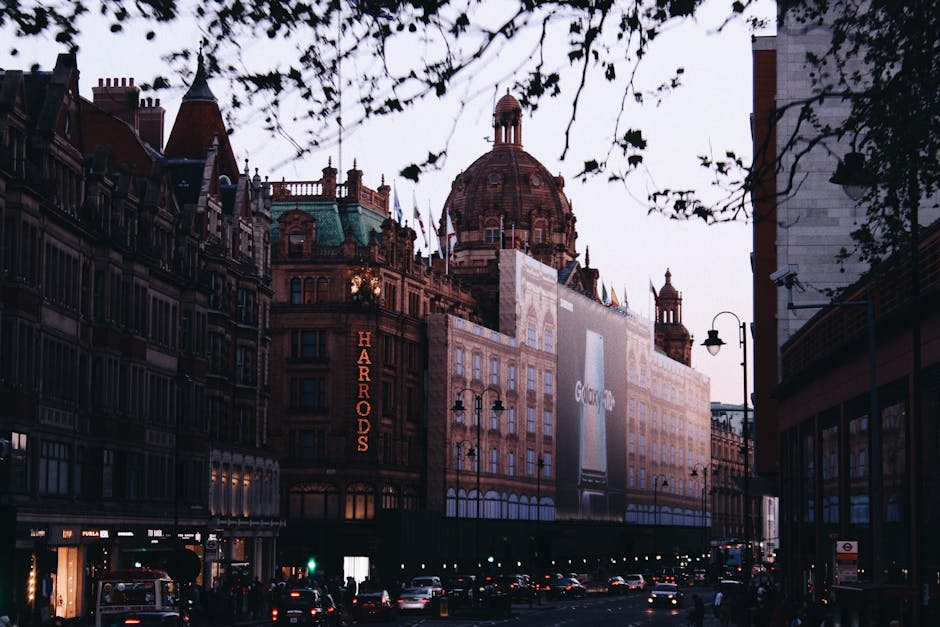 A street scene in a busy urban area during twilight, featuring historic-style buildings with ornate architectural details and a prominent domed structure in the background. The buildings are constructed from brick and stone, with some facades displaying decorative cornices and window frames. A large Harrods sign is visible on one of the buildings on the left, illuminated in red. The street is lined with street lamps and has a steady flow of cars and pedestrians. Overhead, tree branches partially obscure the view, with leaves silhouetted against the dusky sky. To the right, a large billboard with an advertisement for a technology product is mounted on a building, partially covered by scaffolding or protective wrapping. Streetlights cast a warm glow onto the pavement and traffic, creating a contrast with the darker upper areas of the scene. The overall atmosphere captures the lively yet historic character of central London in the evening, with a natural connection to the local environment and the context of urban rubbish removal or private waste management in the area. This scene is associated with a service like Rubbish Removal Knightsbridge, providing alternative waste handling solutions in the vicinity of Harrods and Knightsbridge SW1X.