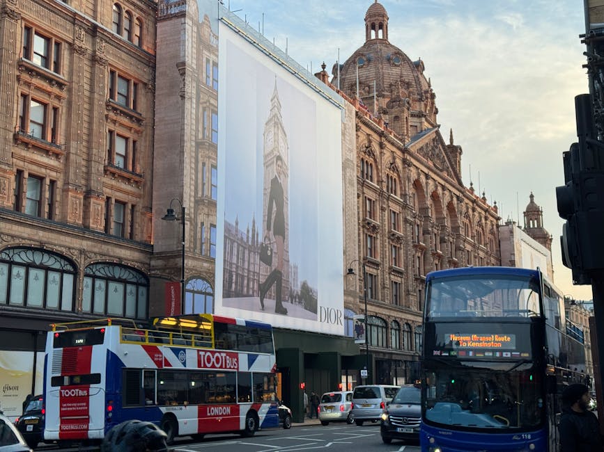A busy city street scene featuring historic, ornate buildings with detailed stone facades and large windows. A prominent billboard displaying a Dior advertisement with an image of a woman walking in front of the Palace of Westminster is mounted on one of the buildings. In the foreground, a double-decker bus marked with 'London' and route information is traveling on the road, accompanied by several smaller vehicles. Pedestrians are visible on the sidewalk, and part of a traffic light can be seen on the right edge of the image. The scene is lit by natural daylight, with a partially cloudy sky overhead, capturing the vibrancy of an urban environment. This image aligns with the context of urban waste management and alternative rubbish collection services, exemplifying a typical cityscape where private rubbish removal might be employed against a backdrop of iconic London architecture and busy transportation hubs.