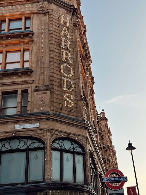 A historic multi-storey building with ornate stone construction featuring detailed architectural embellishments at the corners and window frames. The building's facade displays a vertical sign spelling 'FEARROODS' made from illuminated bulbs or light fixtures, attached along the edge of the structure. The lower part of the building has large, arched window sections with decorative ironwork grilles, and a street sign indicating the intersection of Hans Crescent SW. To the right, a tall street lamp and a classic London Underground sign are visible, with a clear blue sky overhead, suggesting daytime. The scene captures an urban environment typical of a busy city area, aligning with the context of alternative waste handling services like those provided by Rubbish Removal Knightsbridge, which specialize in private rubbish collection and on-site clearance.
