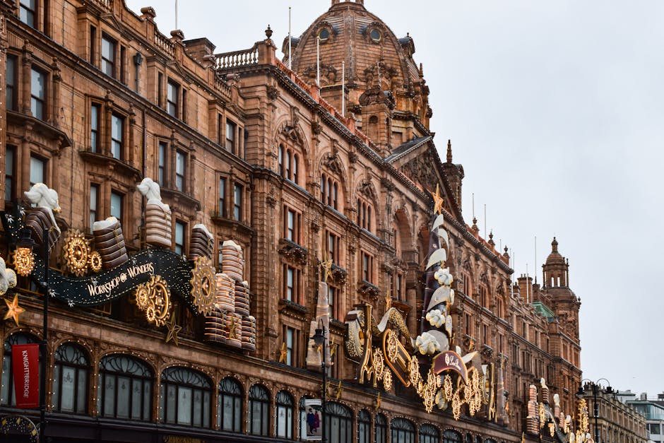 A busy city street scene featuring historic, ornate buildings with detailed stone facades and large windows. A prominent billboard displaying a Dior advertisement with an image of a woman walking in front of the Palace of Westminster is mounted on one of the buildings. In the foreground, a double-decker bus marked with 'London' and route information is traveling on the road, accompanied by several smaller vehicles. Pedestrians are visible on the sidewalk, and part of a traffic light can be seen on the right edge of the image. The scene is lit by natural daylight, with a partially cloudy sky overhead, capturing the vibrancy of an urban environment. This image aligns with the context of urban waste management and alternative rubbish collection services, exemplifying a typical cityscape where private rubbish removal might be employed against a backdrop of iconic London architecture and busy transportation hubs.