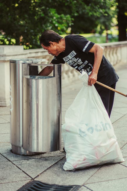 A middle-aged man with dark hair, dressed in a black t-shirt with white text and dark trousers, is seen disposing of rubbish into a large, shiny stainless steel public waste bin on a paved sidewalk. The bin has a cylindrical shape with a rectangular opening at the top, which he is leaning over to access. He holds a long-handled broom in his left hand and a large white plastic bag filled with waste in his right hand. In the background, there is a stone balustrade and lush green foliage, suggesting a park or public garden setting. The scene is illuminated by natural daylight, with shadows cast on the ground. This outdoor environment indicates a typical urban area where rubbish collection involves public waste bins and individual disposal efforts, important in contexts of private rubbish removal or on-site clearance, aligning with services that manage waste outside of regular municipal collection in Knightsbridge.