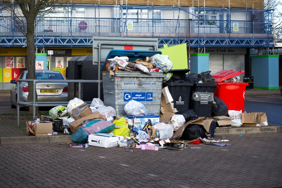 An outdoor scene displaying a large pile of mixed domestic rubbish and waste materials accumulated next to a metal barrier on a paved parking area. The waste includes various cardboard boxes, plastic bags, paper bundles, and discarded packaging, with some flattened and others crumpled. Several black and black-and-white rubbish bags are visible, along with a red waste bin placed to the right of the pile. In the background, a silver car is parked adjacent to the waste, and a multi-storey building with scaffolding and blue safety netting surrounds the upper level. Beneath the scaffolding, shopfront signs and windows are visible, suggesting a commercial or retail environment. The scene is illuminated by daylight, highlighting the textures of the different waste materials and the urban environment surrounding the rubbish. This image illustrates the importance of efficient waste disposal and private rubbish removal services, like those provided by Rubbish Removal Knightsbridge, to manage such waste accumulations properly without reliance solely on municipal collection.