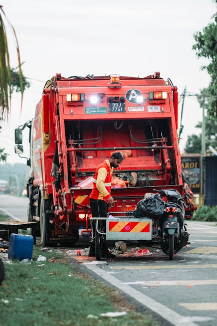 A middle-aged man with dark hair, dressed in a black t-shirt with white text and dark trousers, is seen disposing of rubbish into a large, shiny stainless steel public waste bin on a paved sidewalk. The bin has a cylindrical shape with a rectangular opening at the top, which he is leaning over to access. He holds a long-handled broom in his left hand and a large white plastic bag filled with waste in his right hand. In the background, there is a stone balustrade and lush green foliage, suggesting a park or public garden setting. The scene is illuminated by natural daylight, with shadows cast on the ground. This outdoor environment indicates a typical urban area where rubbish collection involves public waste bins and individual disposal efforts, important in contexts of private rubbish removal or on-site clearance, aligning with services that manage waste outside of regular municipal collection in Knightsbridge.