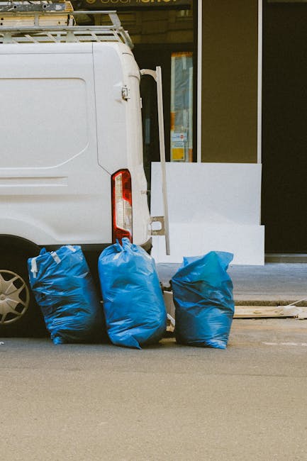 The image shows the rear side of a white commercial van parked on a street, with its tailgate closed. Positioned next to the van are three large blue rubbish bags made of plastic, filled and tied at the top, resting on the pavement. The background includes a building with a large glass window and a white wall section, suggesting an urban environment. The van's rear lights and part of the wheel are visible, while the pavement appears clean and level. The scene depicts an outdoor waste collection or disposal effort, often associated with private rubbish removal services like those offered by Rubbish Removal Knightsbridge, with the blue bags indicating a typical waste clearance process outside commercial or residential premises. The natural lighting reveals details of the bags' textured surface and the van's smooth finish, emphasizing the practical, straightforward arrangement for rubbish clearance or on-site waste handling.